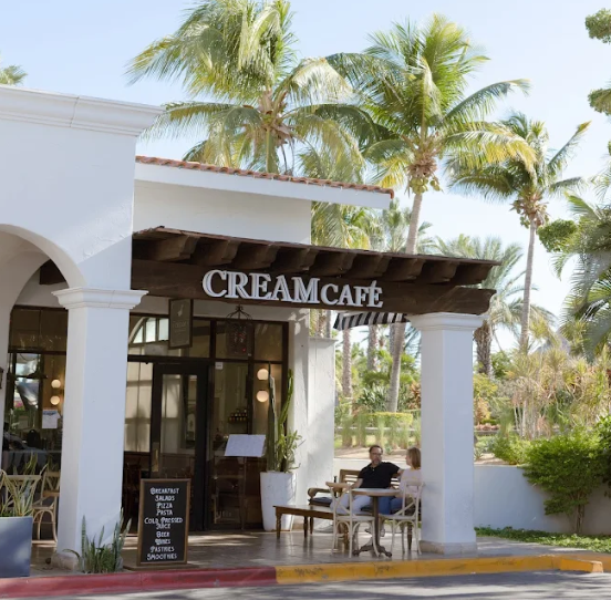 Front entrance of Cream Café in Palmilla, San José del Cabo, with outdoor seating and palm trees in the background on a sunny morning
