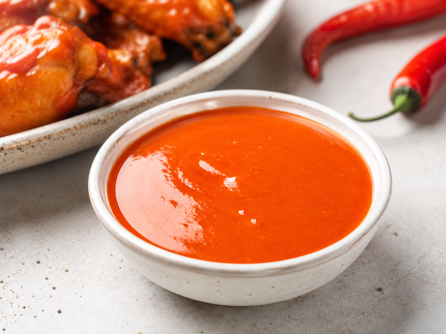 A bowl of buffalo sauce next to buffalo wings and fresh red chili peppers