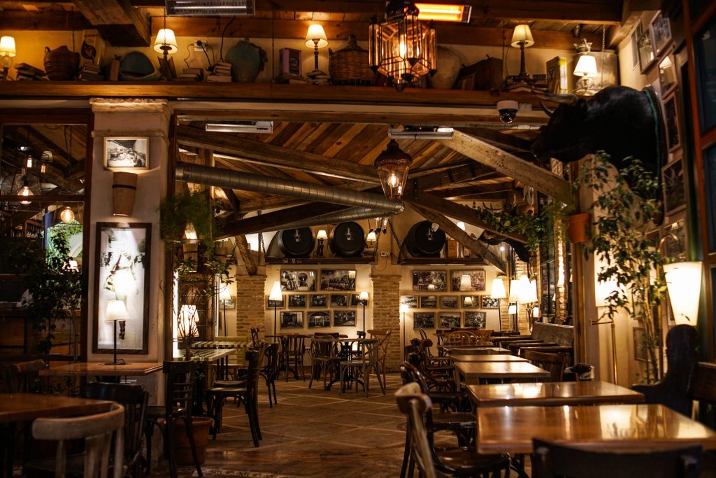 A rustic, warmly lit interior of a restaurant with wooden beams, tables, and chairs. The space features vintag